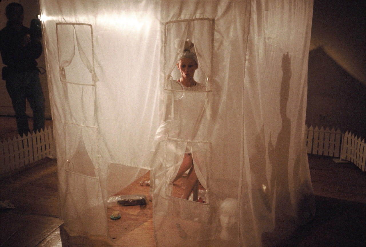 A performer appears to pout, sitting inside a sheer tent in the shape of a 2-storey house