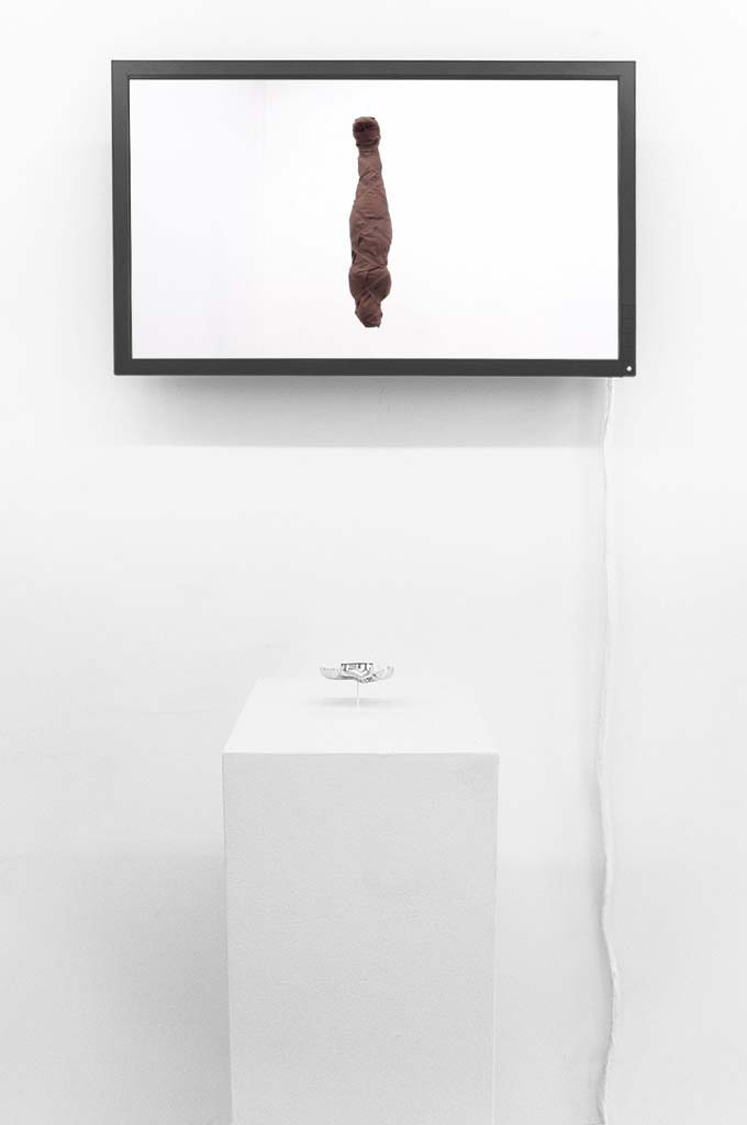 A flatscreen display on a gallery wall, showing an upside-down figure wrapped in dark linen. A plinth in front displays a set of silver false eyelashes.