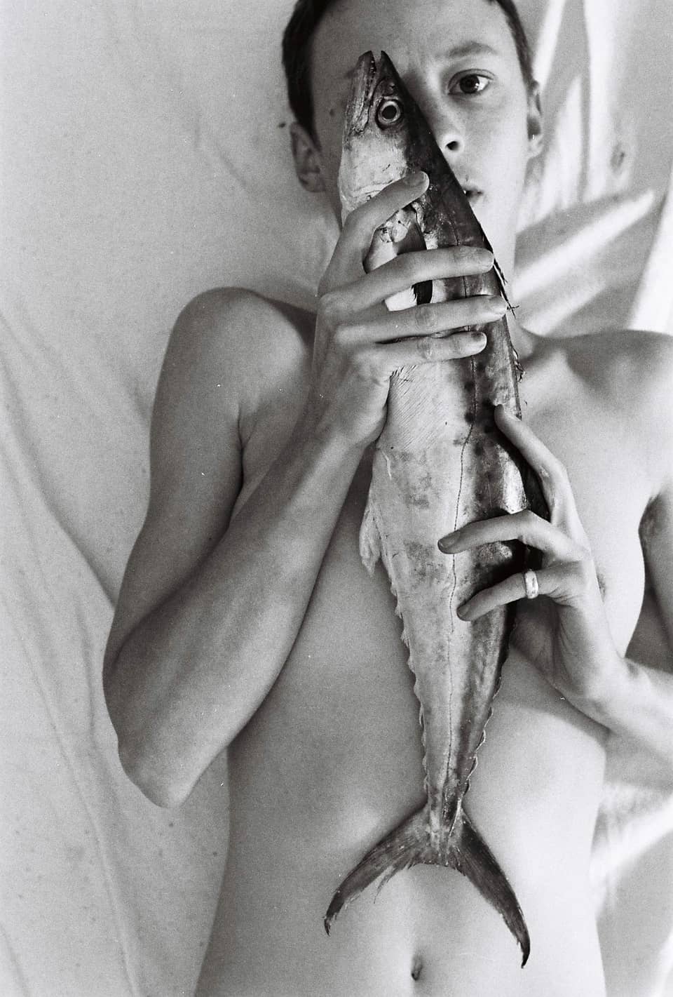 A nude lying on their back, holding a fish. The fish’s eye covers one of theirs.
