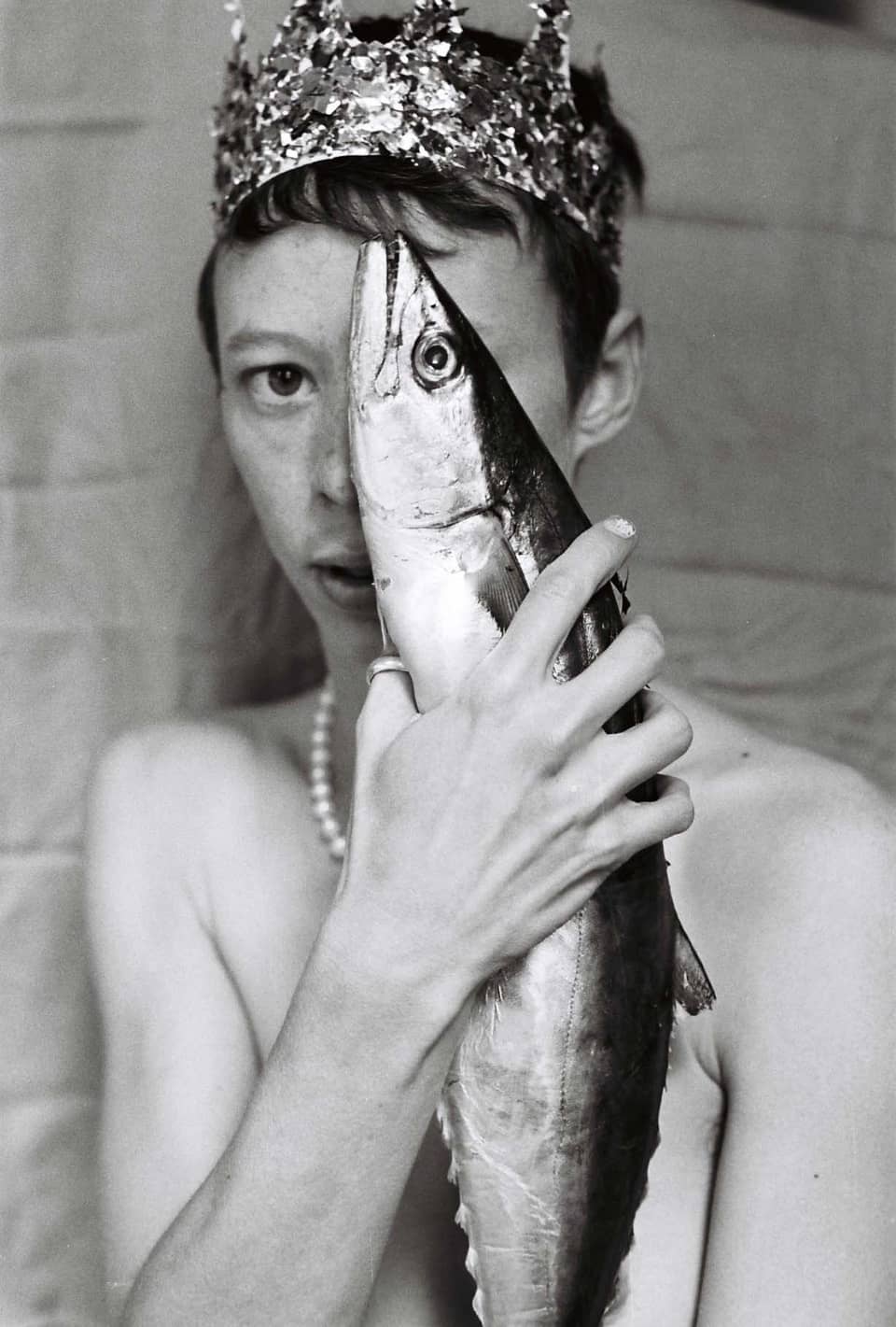 A nude in a crown holding a fish in front of their face. The fish’s eye covers one of theirs.