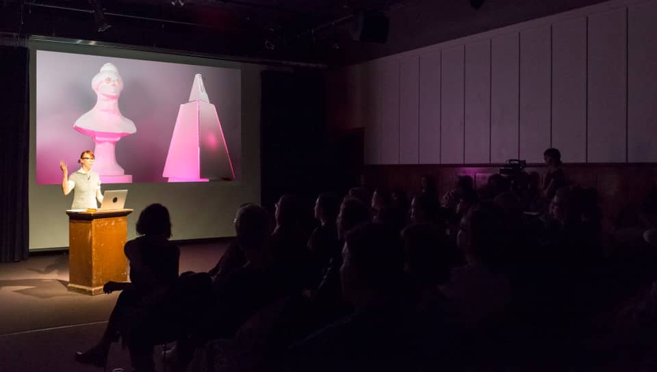 The artist performing, lecture-style, with a large projected image in the background, before a seated audience