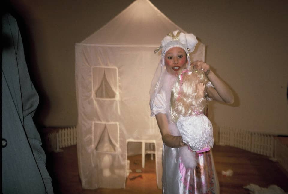 A clown-like figure in white/shiny costume seems to offer a doll to the camera