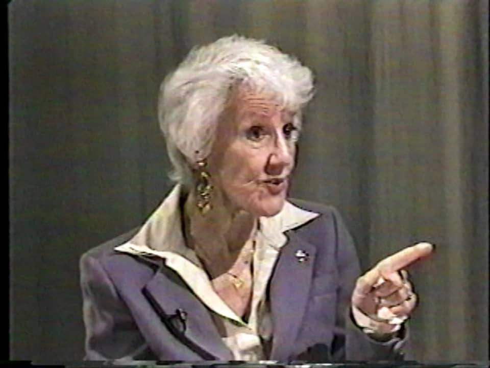 Grey-haired woman in a suit explaining something to someone off-camera