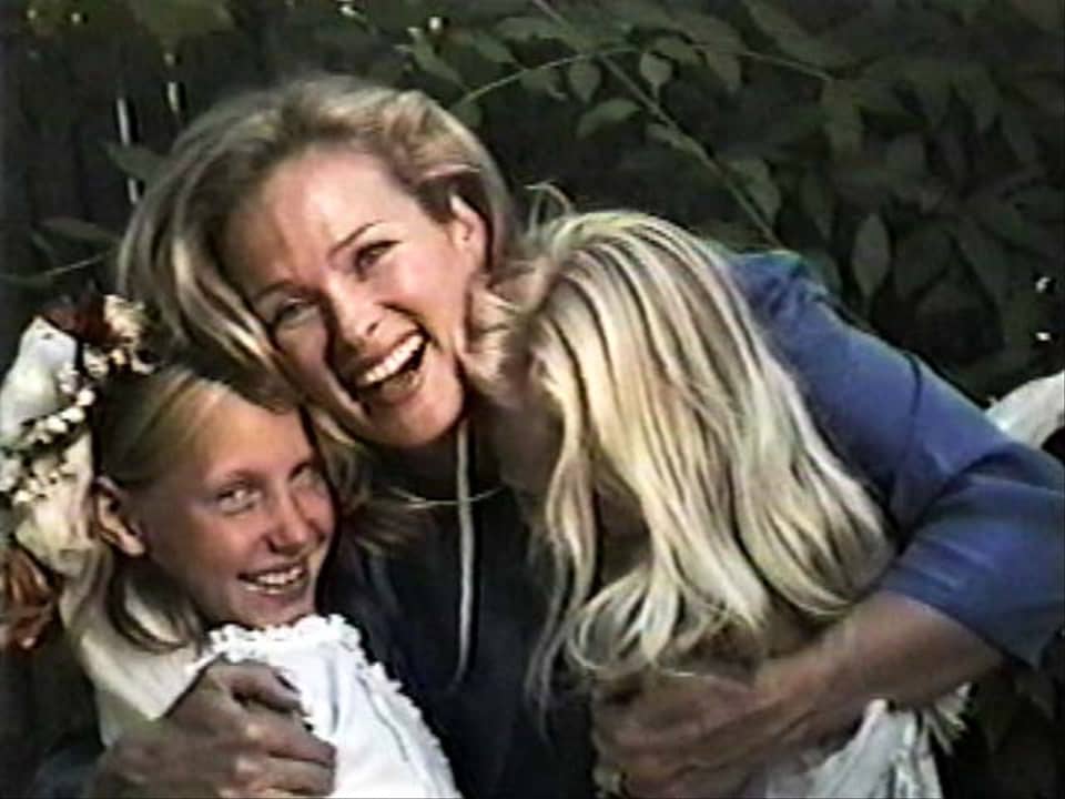 A joyous blonde woman hugs 2 small children while looking into the camera